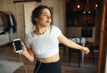 Indoor image of fashionable pretty girl wearing tight fitting top, moving body to rhythm of music, using mobile phone and earphones. People, youth, lifestyle, energy, fun and activity concept