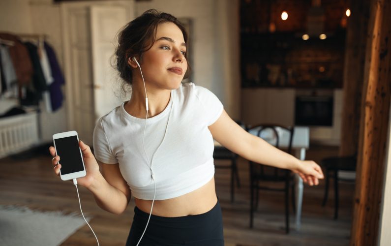 Indoor image of fashionable pretty girl wearing tight fitting top, moving body to rhythm of music, using mobile phone and earphones. People, youth, lifestyle, energy, fun and activity concept
